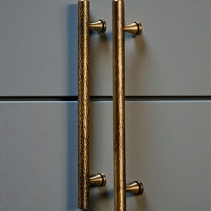 May include: Two bronze-coloured cabinet pulls with a hammered texture, mounted on a grey cabinet door. The pulls are vertical, with a cylindrical bar and rounded end caps. The image is well-lit, highlighting the metallic finish.