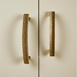 Puede incluir: Dos tiradores de armario de bronce con superficie texturizada montados en una puerta de armario blanca. El tirador de la izquierda es curvo, el de la derecha es recto. La marca "HookzCrafts" está en la esquina superior derecha.