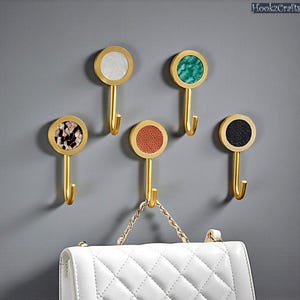 Puede incluir: Cinco ganchos decorativos de pared con tallos dorados y tapas circulares. Las tapas presentan varias texturas y colores, incluyendo blanco, verde, marrón, rojo y negro. Un bolso blanco acolchado cuelga de un gancho.