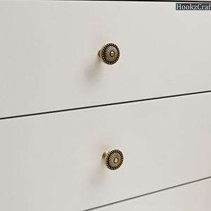 Op de afbeelding: Close-up van een witte kast met twee decoratieve messing knoppen. De knoppen hebben een cirkelvormig ontwerp met een zonnestraalpatroon. De kast heeft drie lades. De knoppen zijn gecentreerd op elke lade.