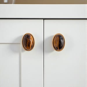 May include: Close-up of two cabinet door knobs. Each knob features an oval-shaped, tiger's eye stone set within a gold-colored metal frame. The knobs are mounted on a white cabinet door. The text "HookzCrafts" is in the top right corner.