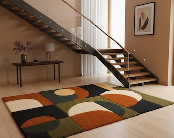 May include: A rectangular area rug with a geometric pattern in shades of green, orange, black, and cream. The rug is placed on a light wood floor, with a staircase and artwork in the background.