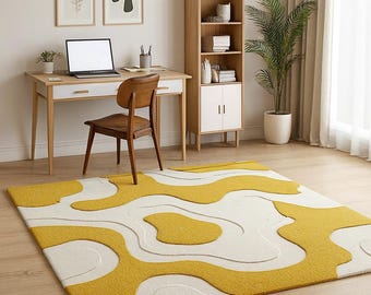 May include: A modern area rug with a cream-colored background and a bold, abstract pattern in shades of yellow. The rug is placed in a room with a wooden desk, chair, and bookshelf. The room also features framed artwork and a potted plant.