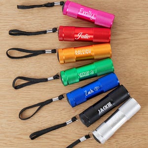 Puede incluir: Una variedad de linternas coloridas, cada una con una correa de muñeca negra. Las linternas son de varios colores, incluyendo rosa, rojo, naranja, verde, azul, negro y plateado. Cada linterna tiene un nombre grabado, como "Emily" y "Julie".