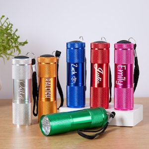 Könnte beinhalten: Eine Auswahl an personalisierten Taschenlampen in Silber, Gold, Blau, Rot und Pink. Jede Lampe trägt einen eingravierten Namen, wie "Zach" und "Julie". Eine grüne Taschenlampe liegt horizontal vor den anderen.