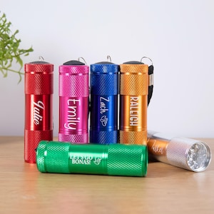 May include: A collection of colorful flashlights in red, pink, blue, orange, green, and silver. Each flashlight has a textured surface and is engraved with a name. The green flashlight has the text "LET'S GO BONAS!"