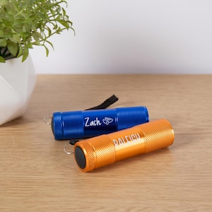 Puede incluir: Dos peque&ntilde;as linternas, una azul y otra naranja, descansan sobre una superficie de madera. La linterna azul tiene el nombre "Zach" impreso, y la linterna naranja tiene el nombre "Raleigh". Ambas linternas tienen una correa de mu&ntilde;eca negra.