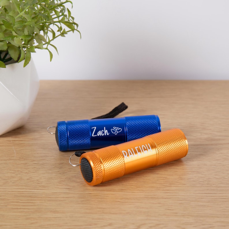 Puede incluir: Dos peque&ntilde;as linternas, una azul y otra naranja, descansan sobre una superficie de madera. La linterna azul tiene el nombre "Zach" impreso, y la linterna naranja tiene el nombre "Raleigh". Ambas linternas tienen una correa de mu&ntilde;eca negra.