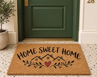 May include: A rectangular tan coir doormat featuring the black text HOME SWEET HOME, a graphic of three small houses, and a red heart centered above a leafy branch design. The mat is placed in front of a dark green door with the number 28.
