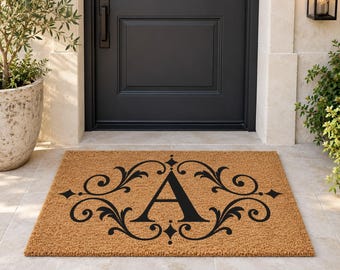 May include: A rectangular tan coir doormat featuring a large black letter A centered within a decorative black scrollwork frame. The design includes symmetrical flourishes and diamond-shaped accents printed on the textured natural fiber surface.