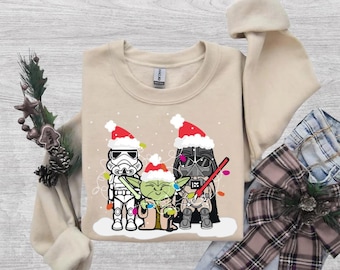 May include: A beige crewneck sweatshirt featuring a graphic print of a Stormtrooper, Grogu, and Darth Vader, each wearing a red and white Santa hat. The characters are tangled in colorful Christmas lights against a white snowy base.