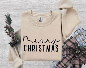 Puede incluir: Una sudadera beige de cuello redondo con el texto negro Merry CHRISTMAS impreso en el frente. La prenda se muestra junto a una rama de pino decorativa con piñas pequeñas y un lazo de tela a cuadros con un detalle de piña.