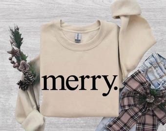 May include: A beige crewneck sweatshirt featuring the word merry. printed in black lowercase letters on the front. The garment is displayed with a decorative pine branch and a plaid ribbon bow, showcasing a simple and clean design aesthetic.