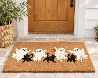 May include: A rectangular tan coir doormat featuring a repeating pattern of white cartoon ghosts and black cats. One ghost is holding a small orange jack-o'-lantern bucket. The design is printed across the center of the textured brown mat.