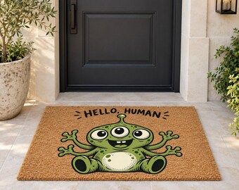 Puede incluir: Un felpudo rectangular de fibra de coco color canela con un alienígena verde de tres ojos, cuatro brazos y vientre claro. El texto HELLO, HUMAN está impreso en negro sobre la ilustración centrada en la parte superior del felpudo.