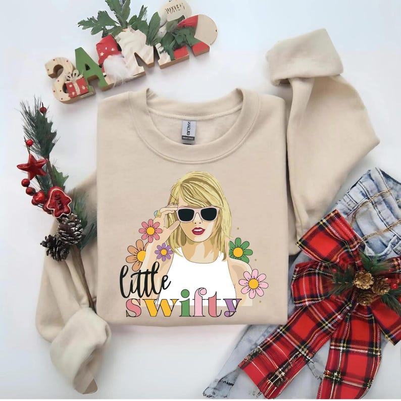 Puede incluir: Una sudadera beige de cuello redondo con una ilustraci&oacute;n gr&aacute;fica de una persona rubia con gafas de sol negras y una camiseta blanca. El dise&ntilde;o incluye flores coloridas y el texto "little swifty" en letras negras y multicolores.