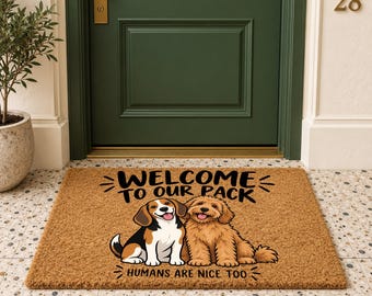 May include: A rectangular tan coir doormat featuring a graphic of a beagle and a golden doodle. The text reads "WELCOME TO OUR PACK" and "HUMANS ARE NICE TOO" in black lettering. The mat is positioned on a tiled floor in front of a green door.