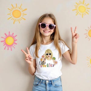 Puede incluir: Una persona joven con una camiseta blanca con un gr&aacute;fico de una persona rubia con gafas de sol y el texto "little swifty". La persona lleva gafas de sol rosas y vaqueros azules. Ilustraciones de sol amarillas y rosas en la pared.