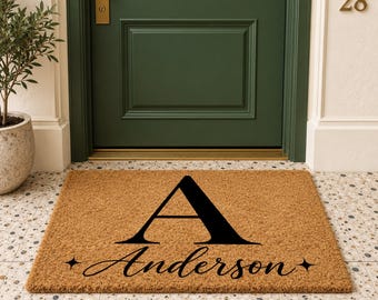 May include: A rectangular tan coir doormat featuring a large black serif letter A centered at the top. Below the letter, the name Anderson is printed in a black script font, flanked by two small four-pointed star icons on each side.