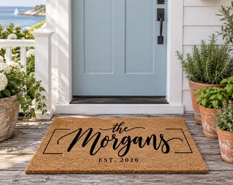 May include: A rectangular tan coir doormat featuring black text that reads The Morgans and EST. 2026, framed by two horizontal lines. The mat is placed on a wooden porch in front of a light blue door with a black handle and white trim.