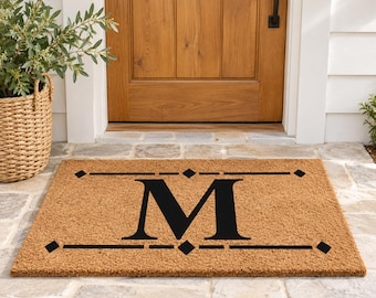 May include: A rectangular tan coir doormat featuring a large black letter M centered in the middle. The letter is framed by horizontal black lines and small black diamond shapes at each corner and the center of the top and bottom borders.