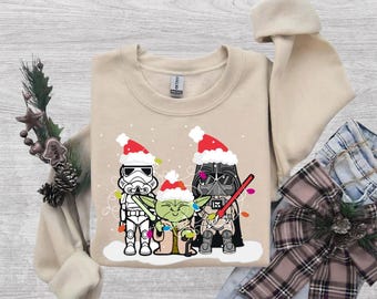 May include: A beige crewneck sweatshirt featuring a graphic print of a Stormtrooper, Yoda, and Darth Vader wearing red and white Santa hats. The characters are tangled in colorful Christmas lights against a backdrop of white snowflakes.