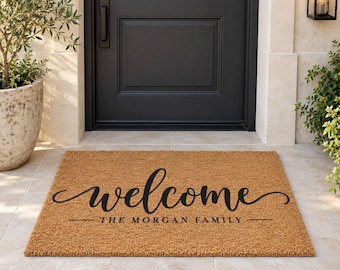May include: A rectangular tan coir doormat featuring the word welcome in a black cursive script font, with the text THE MORGAN FAMILY printed in a smaller, simple black sans-serif font centered directly underneath the word welcome.