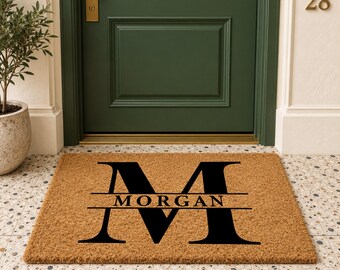 May include: A rectangular tan coir doormat featuring a large black letter M with the name MORGAN printed in black across the center of the letter. The mat is positioned on a tiled floor in front of a dark green door with a gold handle.