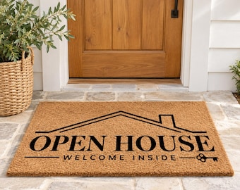 May include: A rectangular tan coir doormat featuring a black graphic of a house outline above the text OPEN HOUSE and the phrase WELCOME INSIDE, finished with a small key icon at the end of the line, centered on the mat surface.