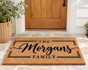 May include: A rectangular tan coir doormat featuring a black border and the text "THE Morgans FAMILY" printed in the center. The word Morgans is written in a large, cursive script font, while "THE" and "FAMILY" are in block letters.
