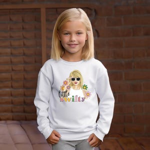 Puede incluir: Una sudadera blanca con un gr&aacute;fico colorido de una mujer con gafas de sol, rodeada de flores, con el texto "little swifty". La sudadera es usada por una ni&ntilde;a.