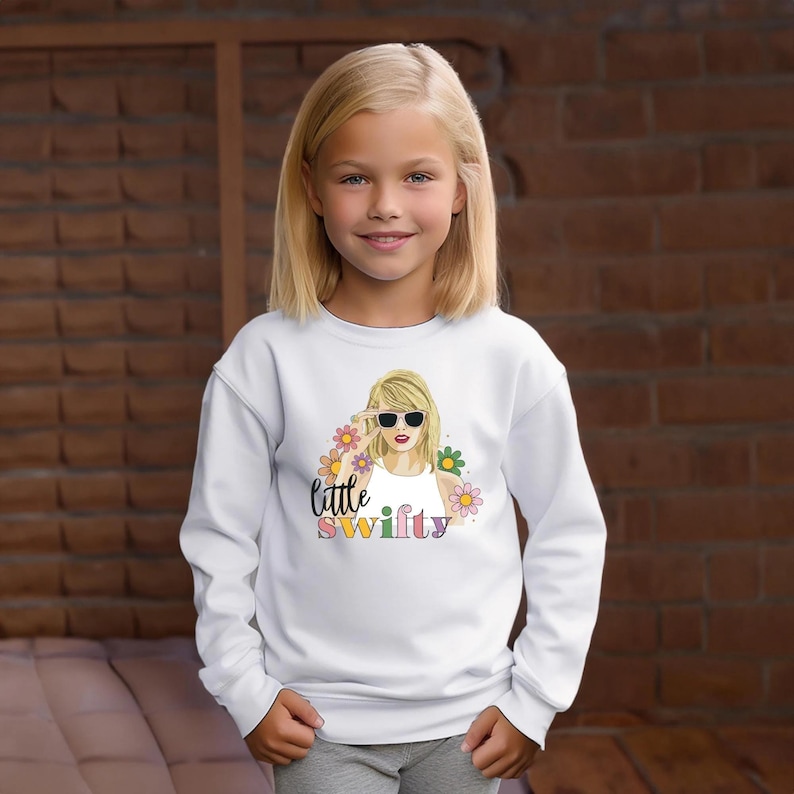 Puede incluir: Una sudadera blanca con un gr&aacute;fico colorido de una mujer con gafas de sol, rodeada de flores, con el texto "little swifty". La sudadera es usada por una ni&ntilde;a.