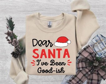May include: A beige crewneck sweatshirt featuring the black and red text Dear SANTA I've Been Good-ish, accompanied by a graphic of a red and white Santa hat. The garment has long sleeves and a ribbed collar, cuffs, and hem.