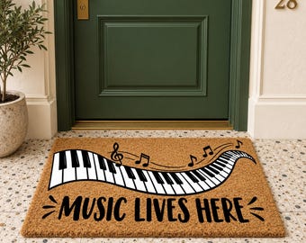 May include: A rectangular tan coir doormat featuring a black and white graphic of a curved piano keyboard with musical notes above it. The text "MUSIC LIVES HERE" is printed in bold black capital letters across the bottom of the mat.