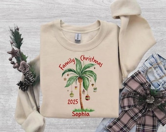 May include: A beige crewneck sweatshirt featuring a graphic of a palm tree decorated with Christmas ornaments. The text reads Family Christmas 2025 and the name Sophia. The design includes green palm fronds and colorful baubles.
