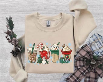 May include: A beige crewneck sweatshirt featuring a graphic print of four holiday-themed beverage cups. The design includes a peppermint-patterned cup, a red mug with a snowman, a green cup with a snowman face, and a red cup with gifts.