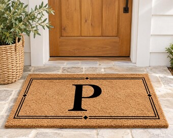 May include: A rectangular tan coir doormat featuring a large black capital letter P centered within a thin black rectangular border. The border design includes small diamond-shaped accents at the center of each side, creating a clean, structured look.
