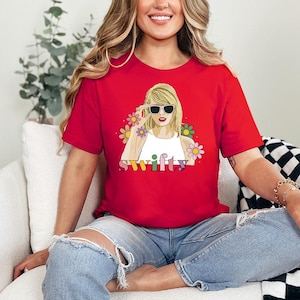 Puede incluir: Camiseta roja con cuello redondo y un gr&aacute;fico de una mujer con gafas de sol, rodeada de flores, con la palabra "Swifty" en letras coloridas. La camiseta es de manga corta.