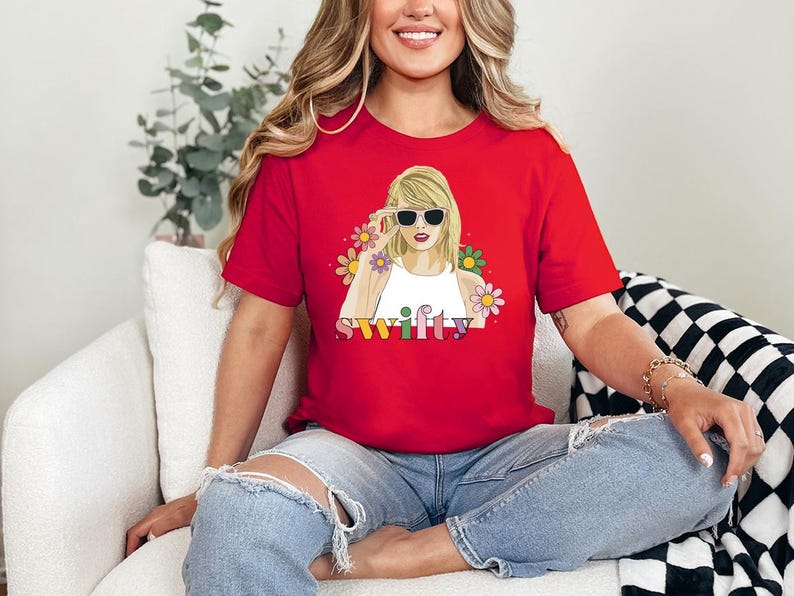Puede incluir: Camiseta roja con cuello redondo y un gr&aacute;fico de una mujer con gafas de sol, rodeada de flores, con la palabra "Swifty" en letras coloridas. La camiseta es de manga corta.