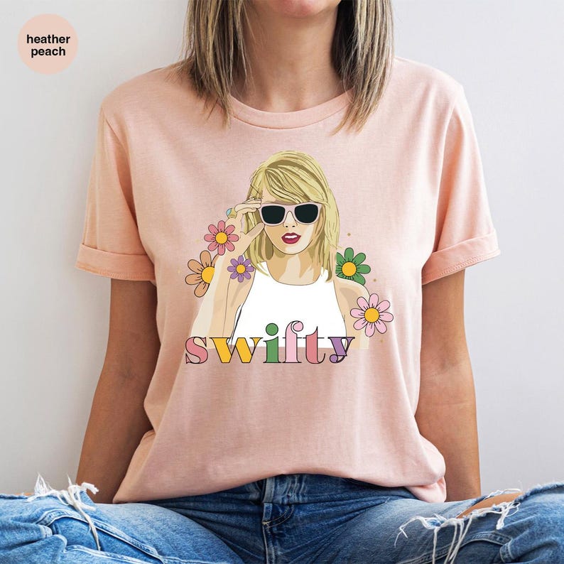 Puede incluir: Camiseta color melocot&oacute;n claro con un gr&aacute;fico de una mujer con gafas de sol, rodeada de flores, con la palabra "Swifty" en letras coloridas. La camiseta tiene mangas enrolladas.