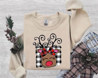 Puede incluir: Una sudadera beige de cuello redondo con un gráfico de una cabeza de reno con astas negras en espiral, un lazo de cuadros rojos y negros y una nariz roja, sobre un fondo cuadrado de cuadros blancos y negros.