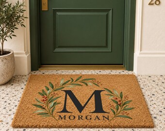 May include: A rectangular tan coir doormat featuring a large black letter M centered within a green leafy wreath with small red berries. The text MORGAN is printed in black capital letters directly below the letter M on the mat.
