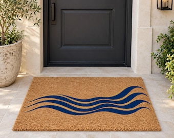 May include: A rectangular tan coir doormat featuring a graphic design of three dark blue horizontal wavy lines that span across the center of the mat, creating a fluid pattern against the natural textured surface of the material.