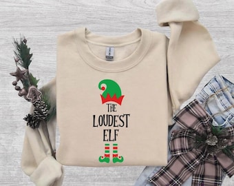 May include: A beige long-sleeve sweatshirt featuring a graphic of a green and red elf hat, the black text "THE LOUDEST ELF", and green and red striped elf legs with green shoes. The garment has a crew neckline and a standard fit.