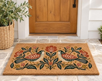 May include: A rectangular tan coir doormat featuring a symmetrical design of two birds facing a central flower, surrounded by dark green leaves, small red berries, and gold star accents, placed on a stone porch in front of a wooden door.