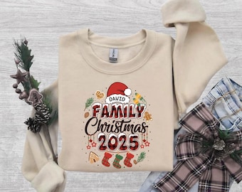 May include: A beige crewneck sweatshirt featuring a graphic with the text DAVID FAMILY Christmas 2025. The design includes a red Santa hat, red and black plaid lettering, three hanging Christmas stockings, and small holiday icons.