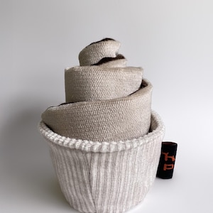 Könnte beinhalten: Eine beige gewebte Stoffkrawatte, gerollt und in einem passenden gerippten Strickbehälter verschachtelt. Die Innenseite der Krawatte ist dunkler braun. Ein kleines schwarzes Etikett mit orangefarbenem Text befindet sich neben dem Behälter.