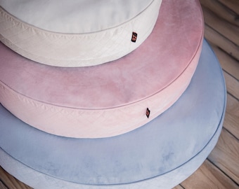 May include: Three circular pet beds in pastel shades of blue, pink, and white, stacked on top of each other. Each bed features a quilted design and a small label. The beds are on a wooden floor.