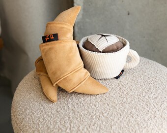 May include: A plush croissant and a coffee cup toy set. The croissant is tan with a black tag that has orange text. The coffee cup is white with a ribbed texture and a brown top with a white and brown striped square.