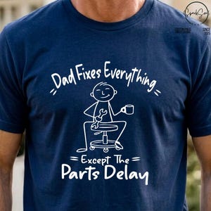 Könnte beinhalten: Ein marineblaues Kurzarm-T-Shirt mit einer weißen Grafik eines Strichmännchens, das auf einem Hocker sitzt und einen Schraubenschlüssel sowie eine Tasse hält. Der Text lautet: Dad Fixes Everything Except The Parts Delay. Das Motiv ist mittig.